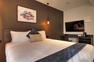 Una habitación de hotel moderna con una cama grande, un televisor de pantalla plana, un escritorio con cafetera y una pieza de arte único en la pared.
