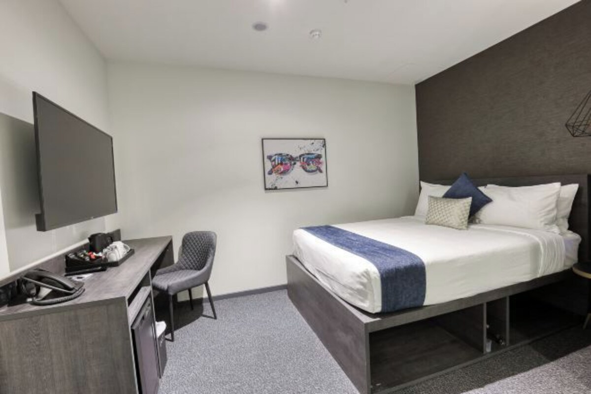 A modern hotel room with a bed, a desk, a chair, and a flat-screen TV.