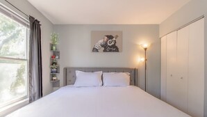 Et soveværelse med en stor seng, et sengebord med en lampe, et vindue med gardiner og en vægmonteret hylde med planter.