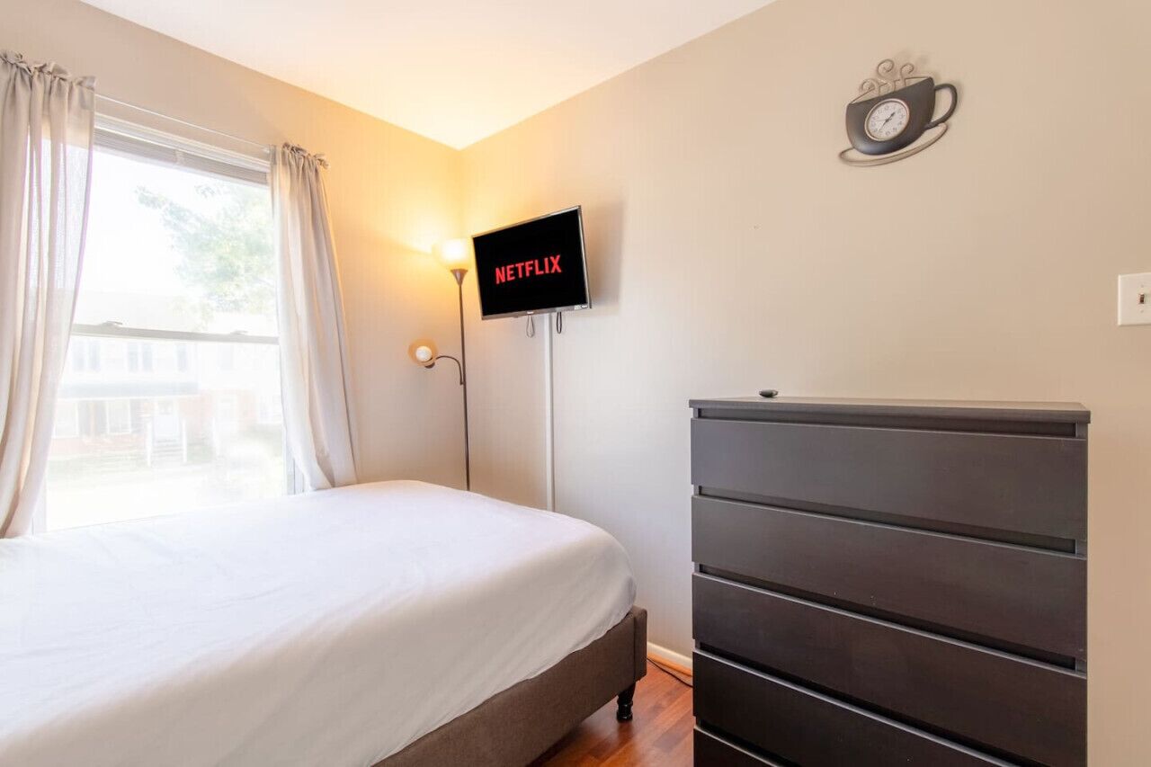 Un dormitorio con una cama, un aparador, un televisor montado en la pared que muestra 'NETFLIX' y un reloj de pared.