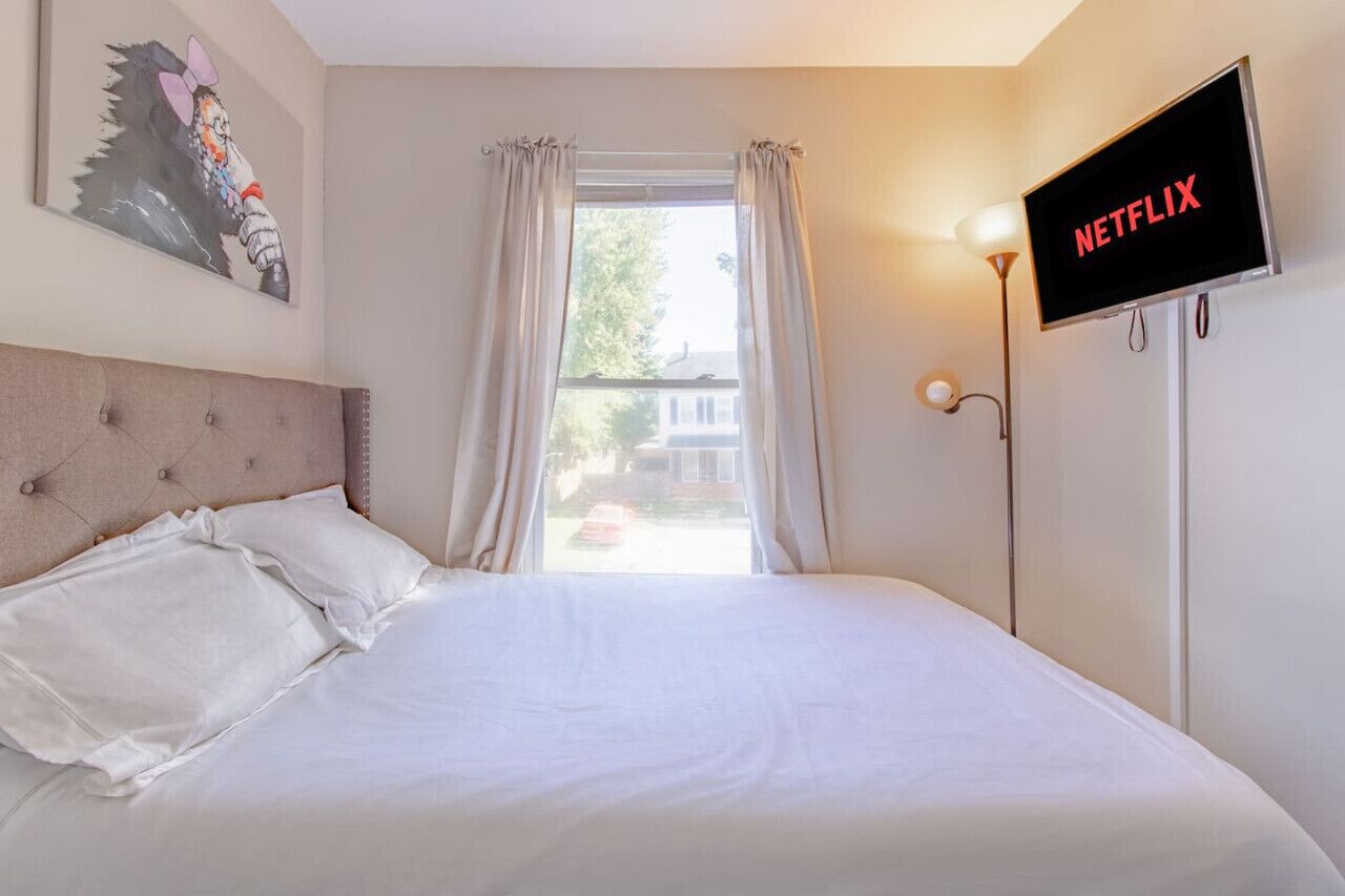 Un dormitorio con cama, ventana con cortinas, televisor montado en la pared mostrando 'NETFLIX' y un cuadro en la pared.