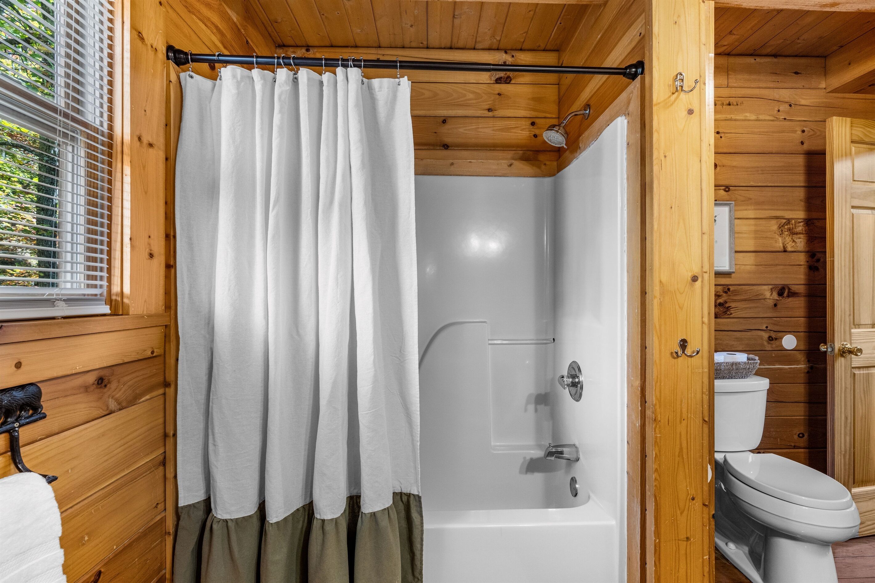 A wooden cabin bathroom with a shower, toilet, and window with blinds.