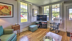 Una habitación con piso de madera, televisor, mecedora, una mesita y ventana con vista a árboles y edificios.