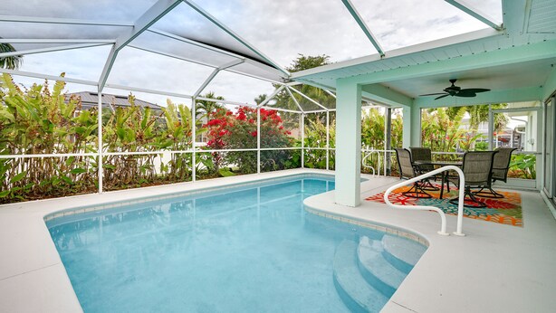 Una piscina interior con techo de cristal, rodeada de vegetación y un área de patio con sillas y una mesa.