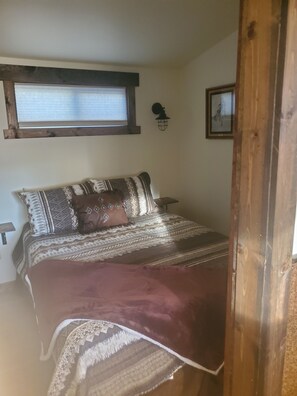 A bedroom with a bed, a window, a wall-mounted light fixture, and a framed picture. - Three Forks Saddlery's Stable (Three Forks)