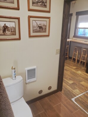 A bathroom with a toilet, a wall-mounted heater, and framed pictures on the wall. - Three Forks Saddlery's Stable (Three Forks)