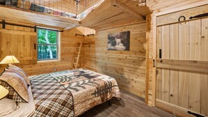 A wooden cabin bedroom with a loft area, a bed with a plaid bedspread, a window with a view of trees, and a wooden door.