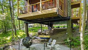 A wooden cabin with a stone foundation, a swing chair, a fire pit, and a view of the surrounding forest.