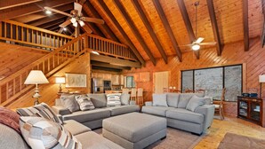 A spacious living room with a high ceiling, wooden beams, a staircase, and a kitchen area.