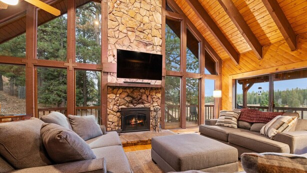 A spacious living room with a stone fireplace, large windows, and wooden beams.