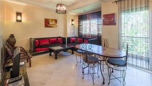 A living room with a sofa, a dining table with chairs, a chandelier, and a large window with blinds. - Residence Bouroud Hivernage 5376 (Marrakech)