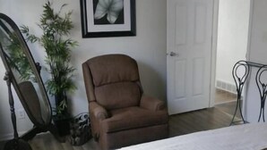 A bedroom with a bed, a brown chair, a framed picture on the wall, and a door. - Cute and Cozy (Rockford)