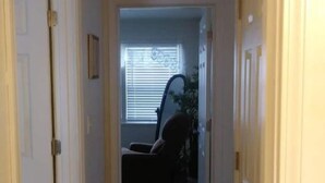 A narrow hallway with a dark floor leading to a room with a window and a chair. - Cute and Cozy (Rockford)
