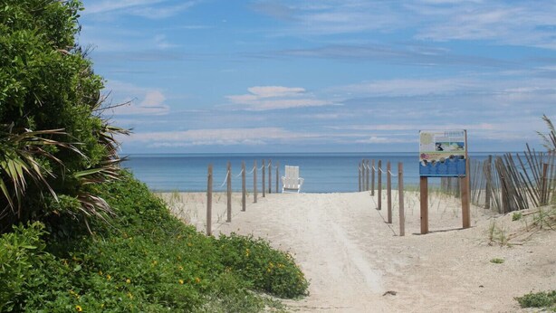 Un sentier de sable qui longe la plage et mène à l’océan, avec une clôture en bois et une chaise blanche.