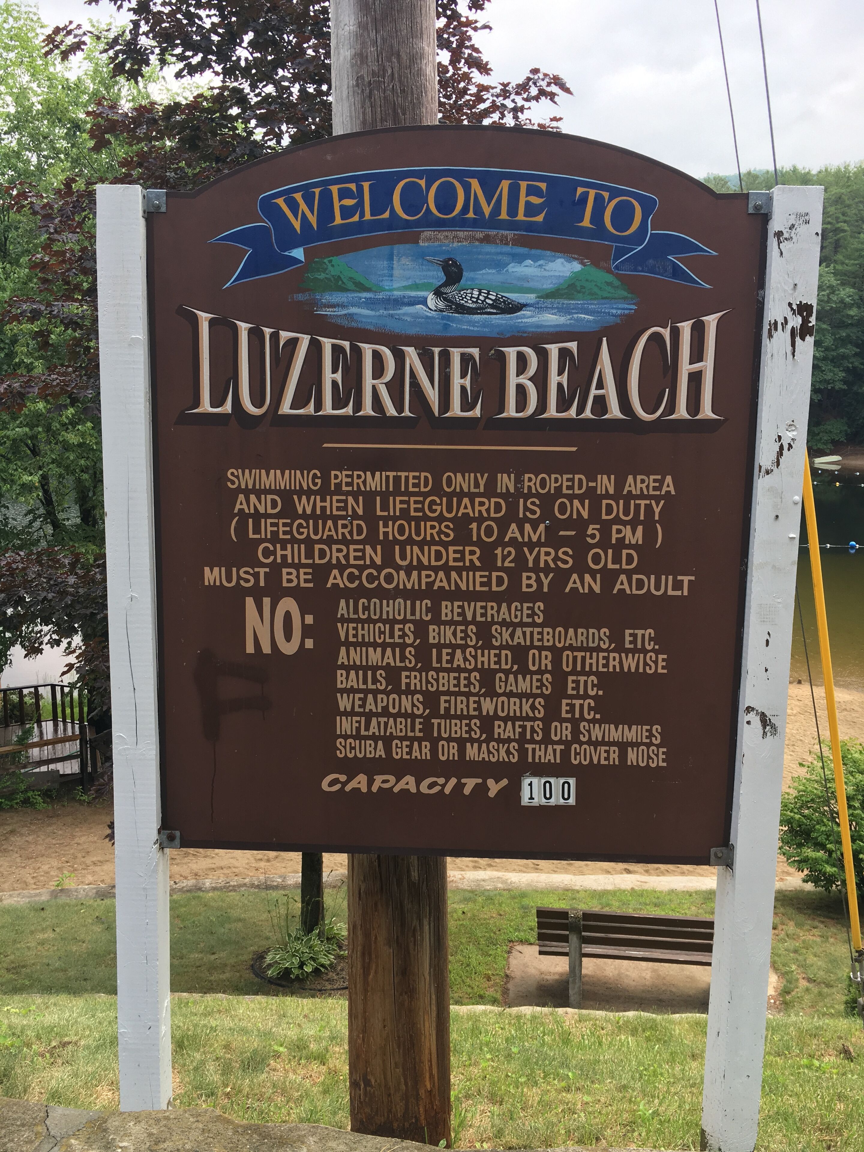 En välkomstskylt för Luzerne Beach med information om badregler och maximalt antal besökare: 100.