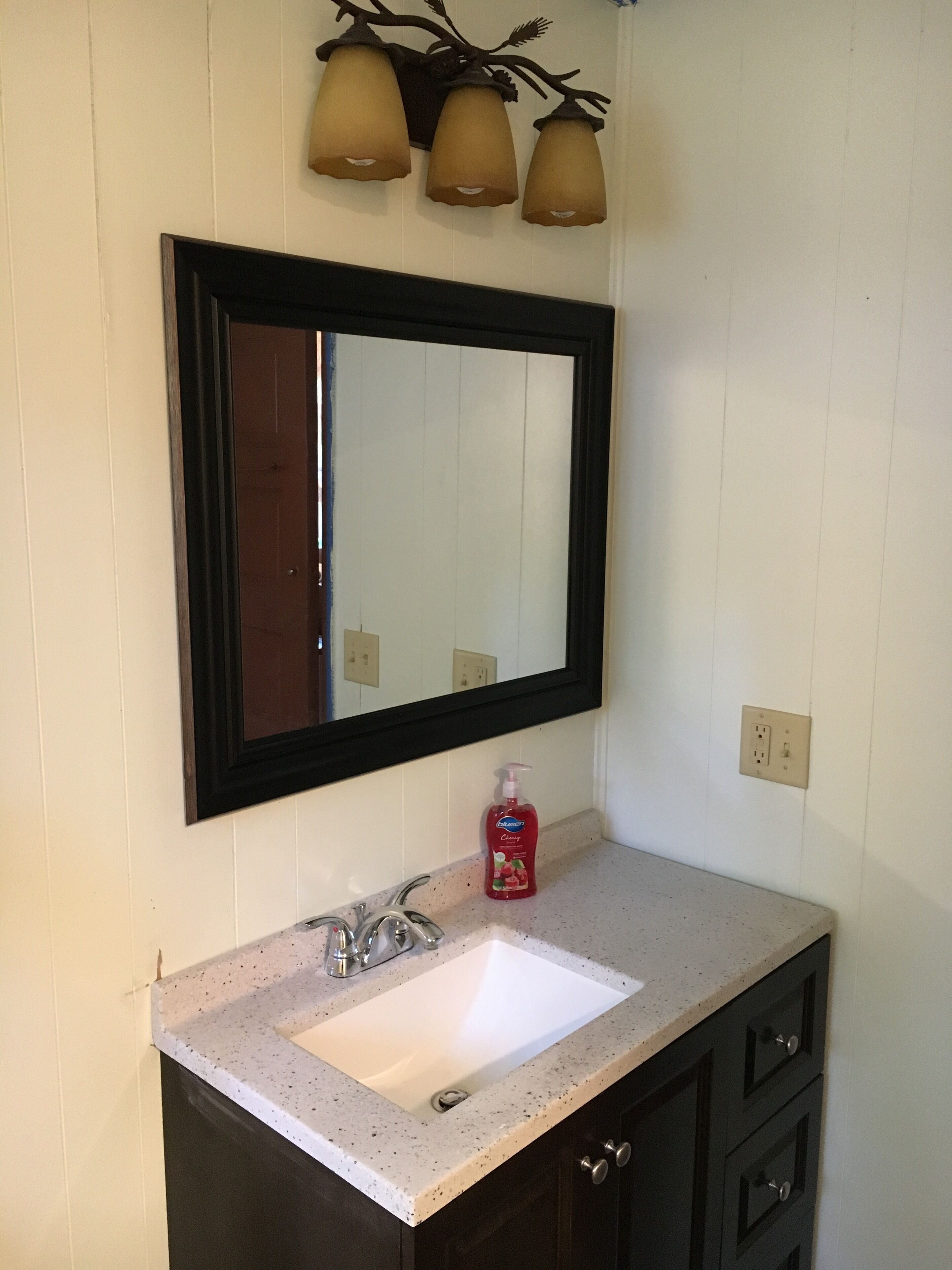A bathroom with a white sink, dark cabinets, a mirror with a black frame, and three wall-mounted lights.