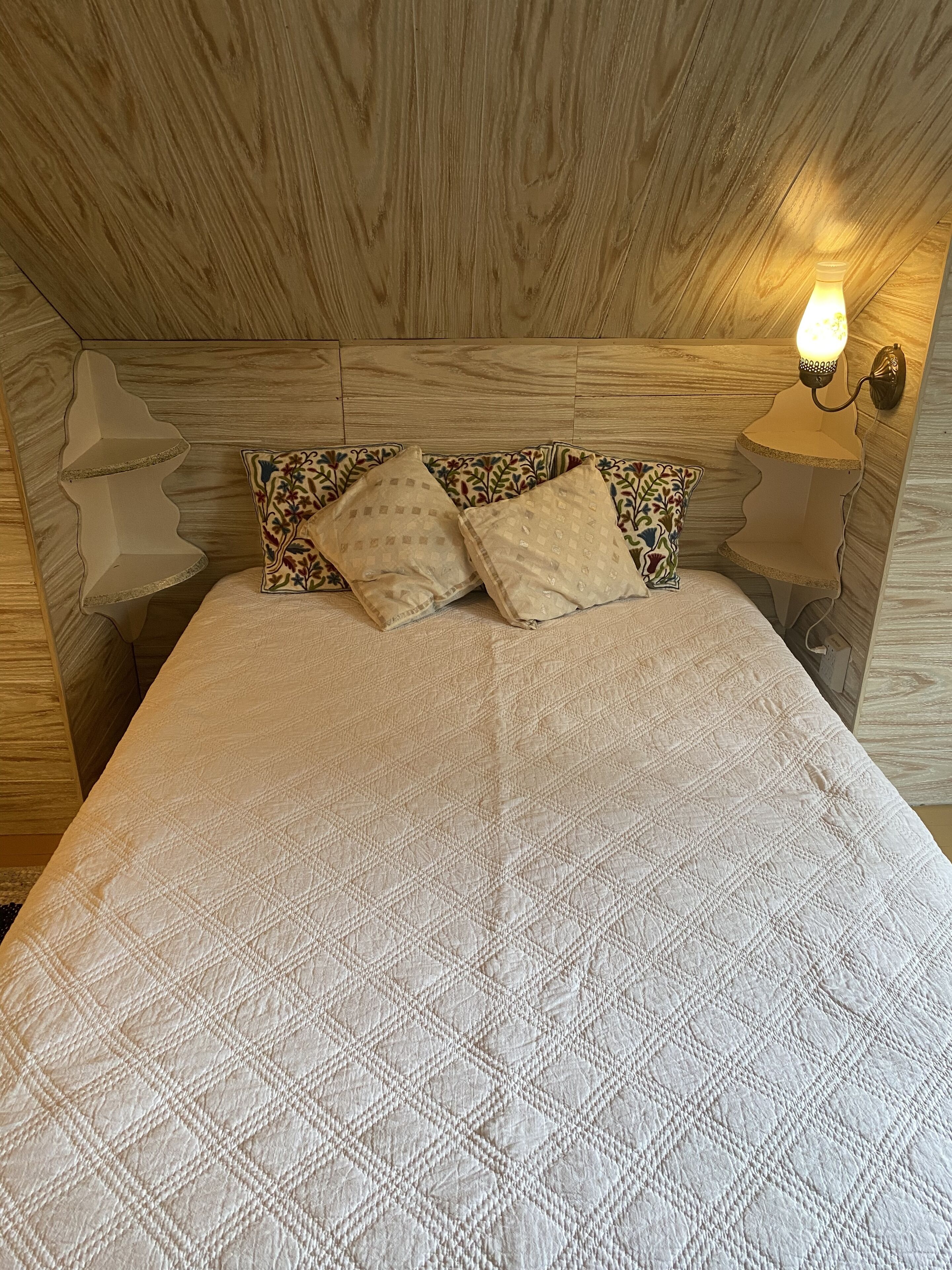 A bed with a quilted white cover, two patterned pillows, and a wall-mounted light fixture.