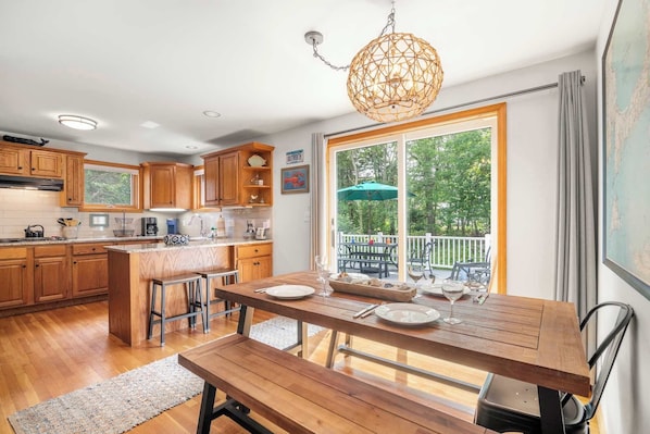 A kitchen with wooden cabinets, a dining table with benches, and a large window with a view of the outdoors. - Central AC Close to Nauset Beach! (Orleans)