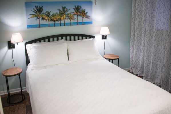 A neatly made bed with two bedside tables, each with a lamp, in a room with a light blue wall and a tropical-themed painting above the bed.