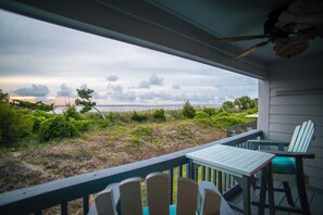 A balcony with a view of the beach, a table, two chairs, and a ceiling fan.