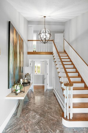 A spacious hallway with a marble floor, a wooden staircase, and a large abstract painting on the wall. - NEW HOT TUB||sleeps 10|4BR 3Ba|firepit|pingpong table|New River Gorge NP (Fayetteville)