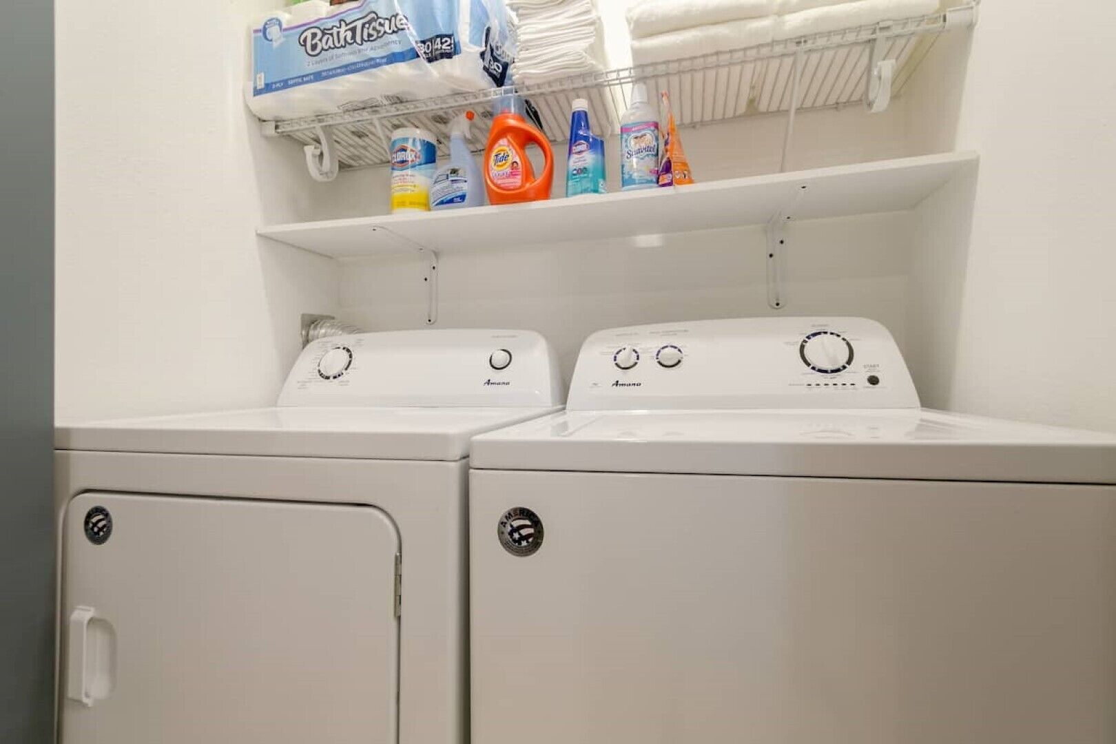 A laundry area with two front-loading washing machines and a shelf above containing various laundry detergents.