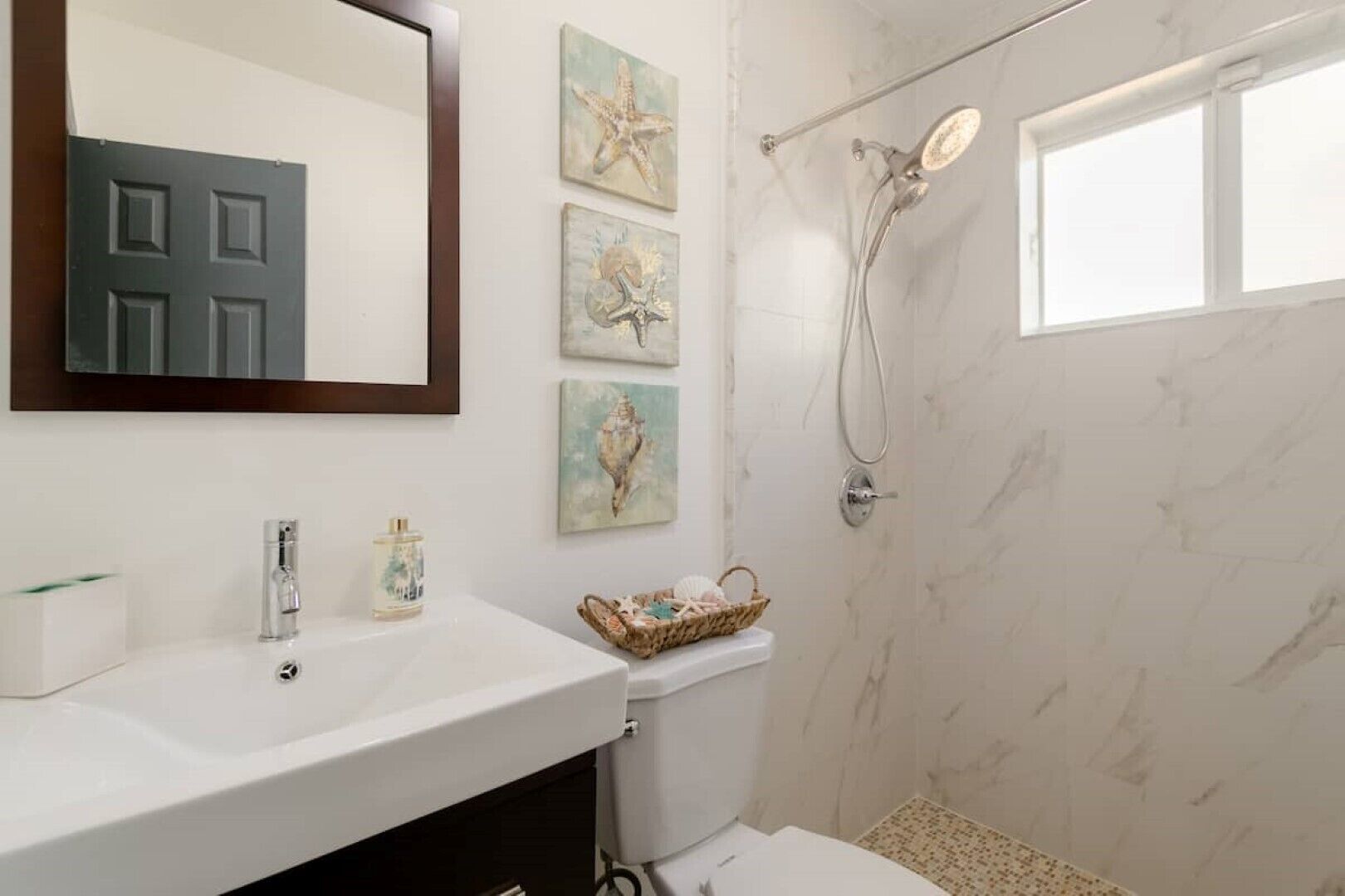 A bathroom with a white sink, a toilet, a shower with a handheld showerhead, and three framed artworks on the wall.