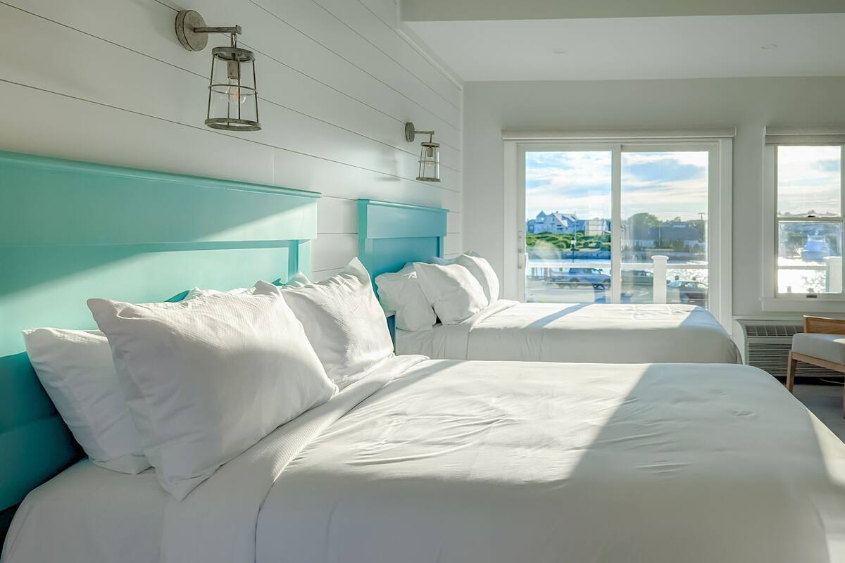 Habitación de hotel con dos camas, cabeceros verde azulado, ropa de cama blanca y vistas a la ciudad.