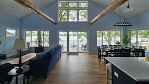 A spacious living area with a high ceiling, wooden beams, a blue sofa, a dining table, and large windows offering views of a lake. - Pearl Beach:  Dive into Summer Fun at Pearl Beach – Swim, Kayak & Relax! (Greenville)