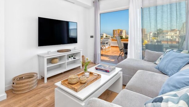 A modern living room with a flat-screen TV, white furniture, a wooden coffee table, and a view of the city through large windows. - Lola Playa by Interhome (Benalmádena Costa)