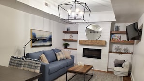 A modern living room with a blue sofa, a wooden coffee table, a TV, and a fireplace. - Clark Suite - Millpond Inn (Clarkston)