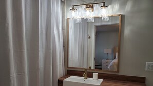 A bathroom with a wooden vanity, a white sink, and a mirror with a light fixture above it. - Clark Suite - Millpond Inn (Clarkston)