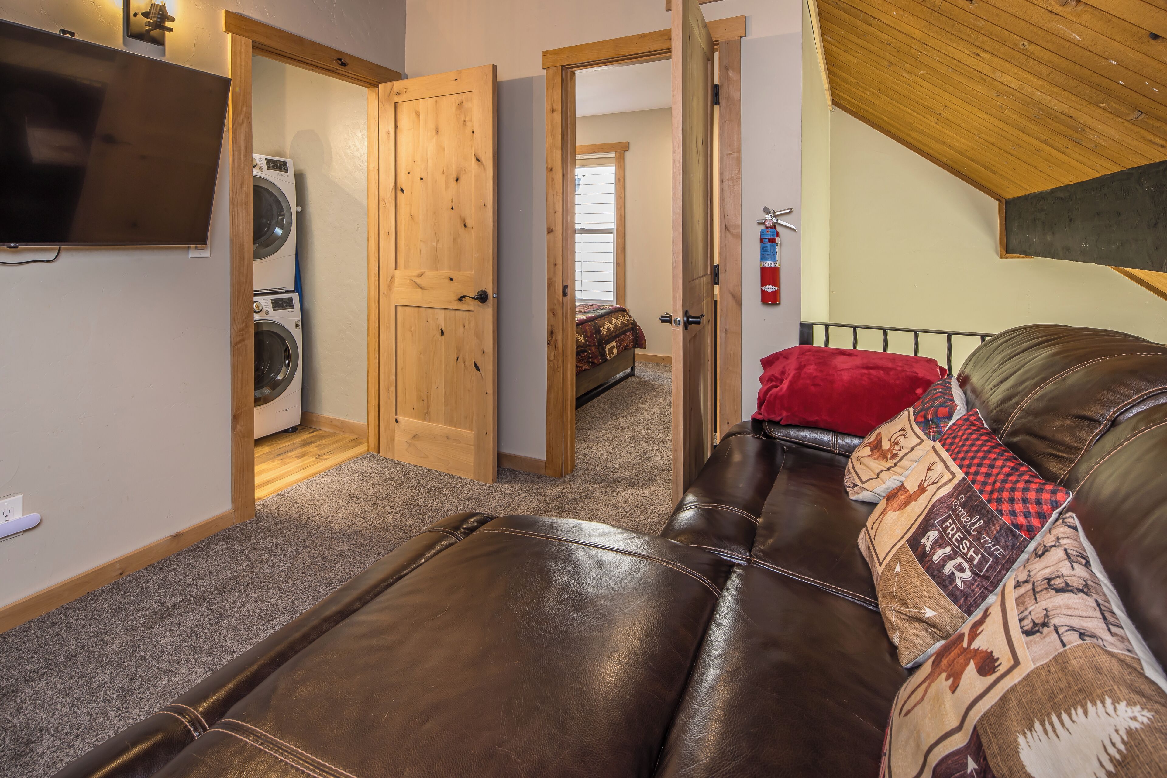 A room with a brown leather couch, a television mounted on the wall, and a wooden door leading to another room.