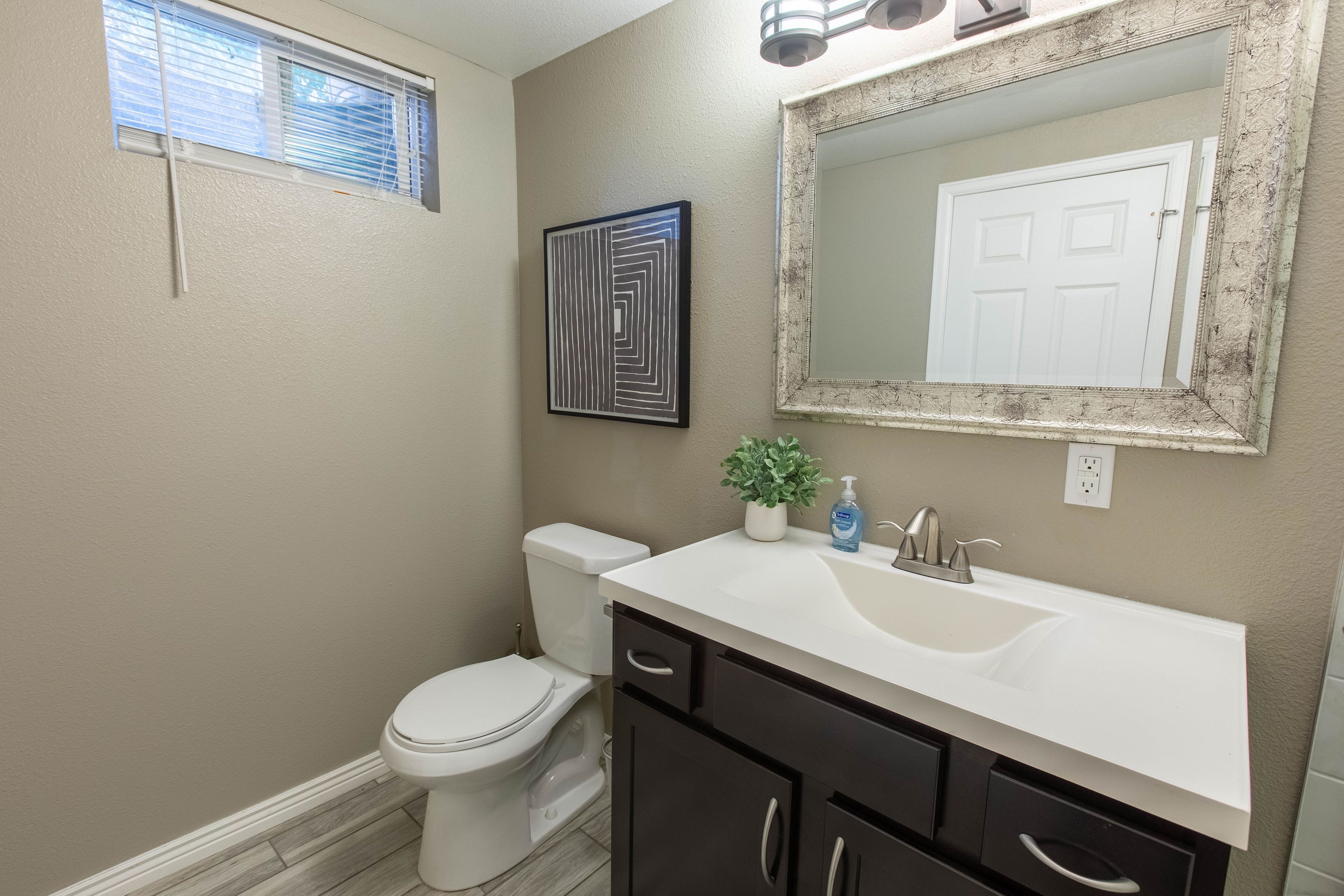 Une salle de bain avec des toilettes blanches, un lavabo surmonté d’un miroir et un tableau encadré accroché au mur.