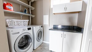 A laundry room with two front-loading washing machines, a white laundry basket, and shelves with cleaning supplies. - NEW Renovated Guest Suite | 30 min to DIA & DEN (Brighton)