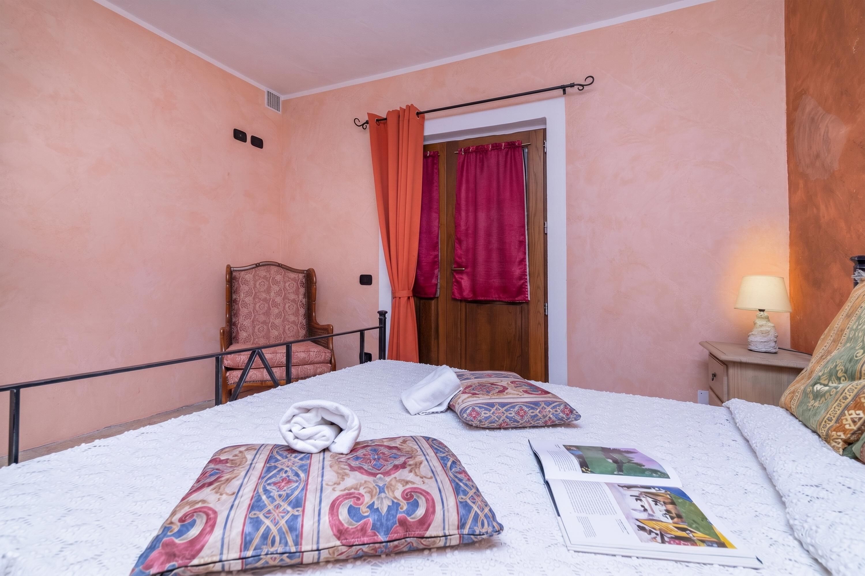 A bedroom with a bed, a chair, a nightstand with a lamp, and a door with curtains.