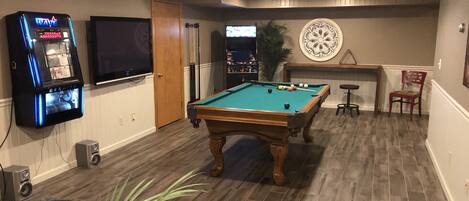 A pool table in a room with wooden flooring, a television mounted on the wall, and a dartboard on the wall.