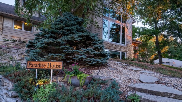 A house named 'Paradise House' with a stone wall, a large coniferous tree, and a well-maintained garden with various plants and a wooden sign.