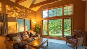 A spacious living room with a leather sofa, a patterned armchair, a wooden coffee table, and large windows offering a view of the outdoors.