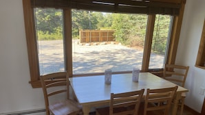 A dining area with a wooden table and chairs, large windows with a view of a wooden deck and trees. - Duck Cove Cottage overlooking beautiful Blue Hill Bay (Bernard)