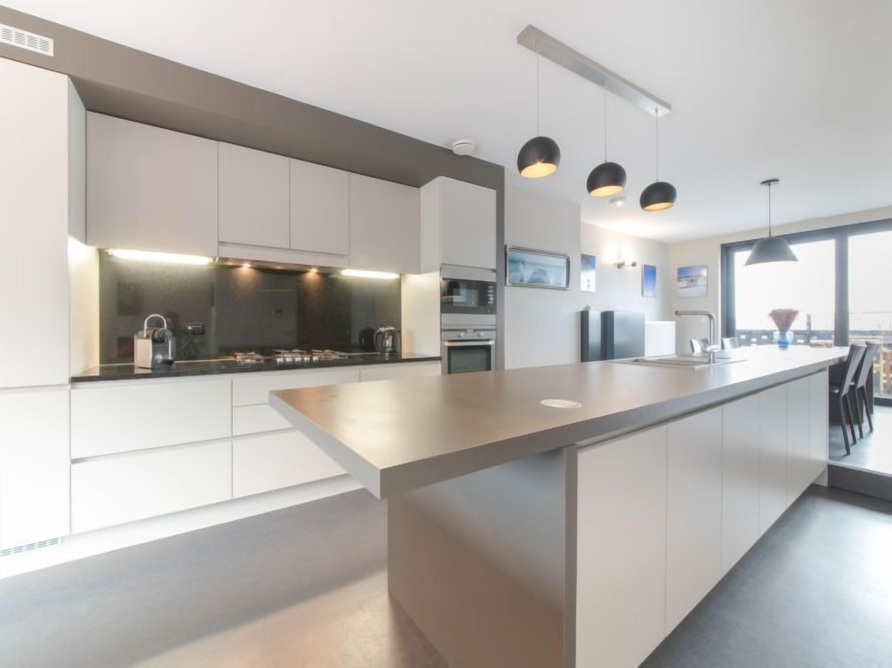 A modern kitchen with a central island, built-in appliances, pendant lighting, and a view of the city through large windows.