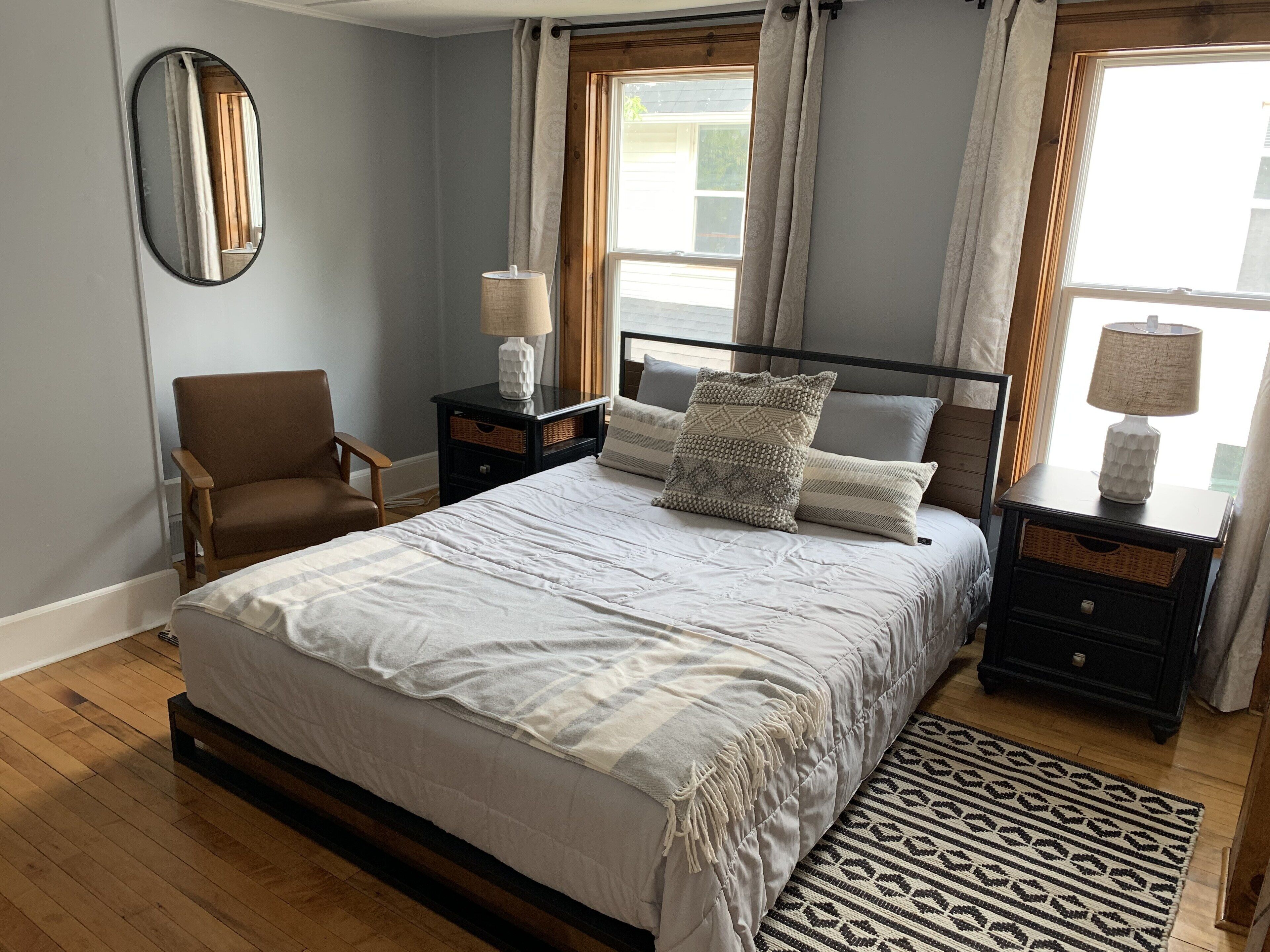 Ein Schlafzimmer mit einem großen Bett, zwei Nachttischen, einem Sessel, einem Spiegel und einem Fenster mit Vorhängen.