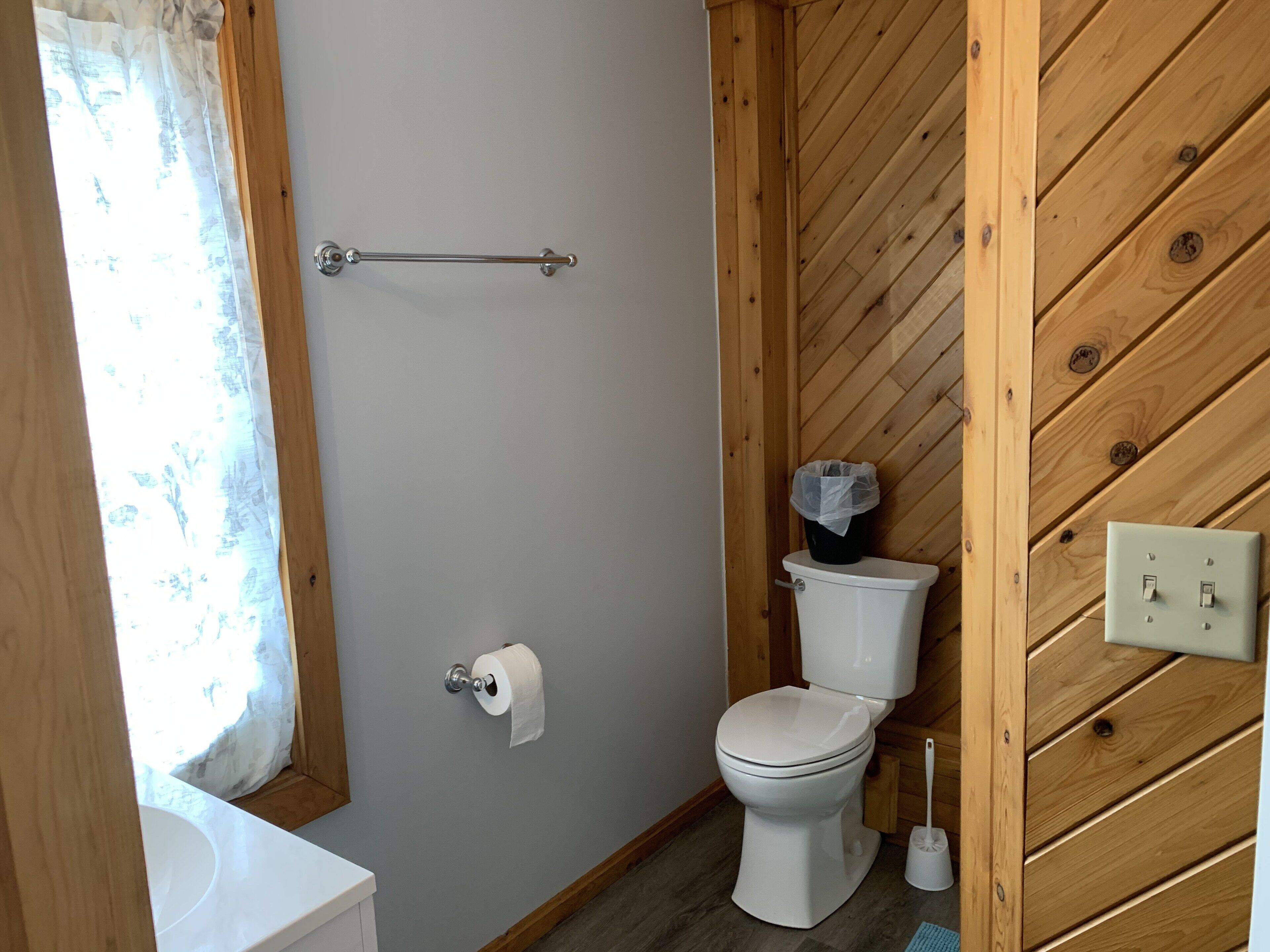 Een badkamer met een houten wand, een wit toilet, een rol toiletpapier, een handdroger en een raam met gordijn.