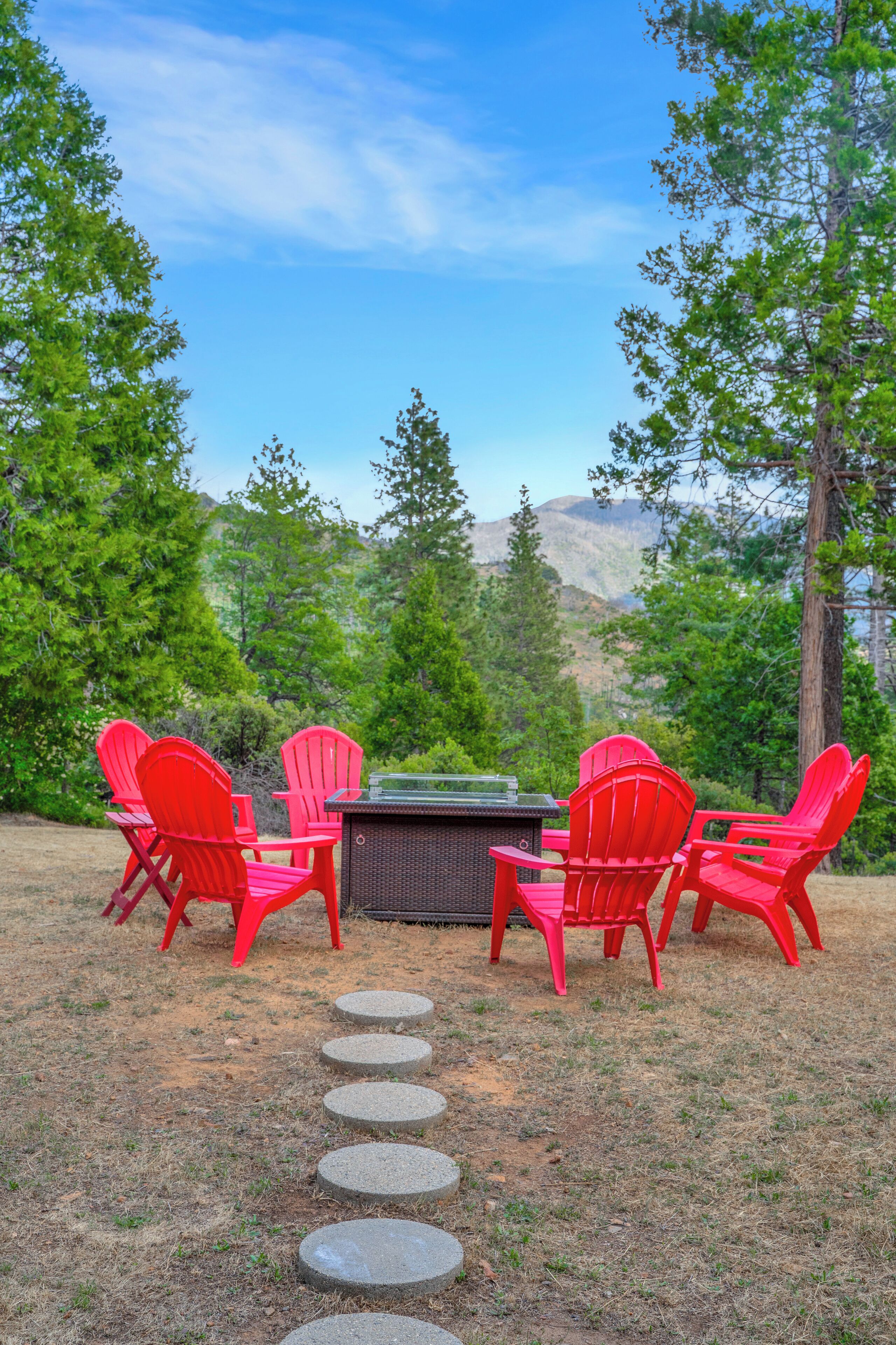 Un espacio al aire libre con sillas Adirondack rojas y una mesa, rodeado por un paisaje natural con árboles y vistas a montañas.