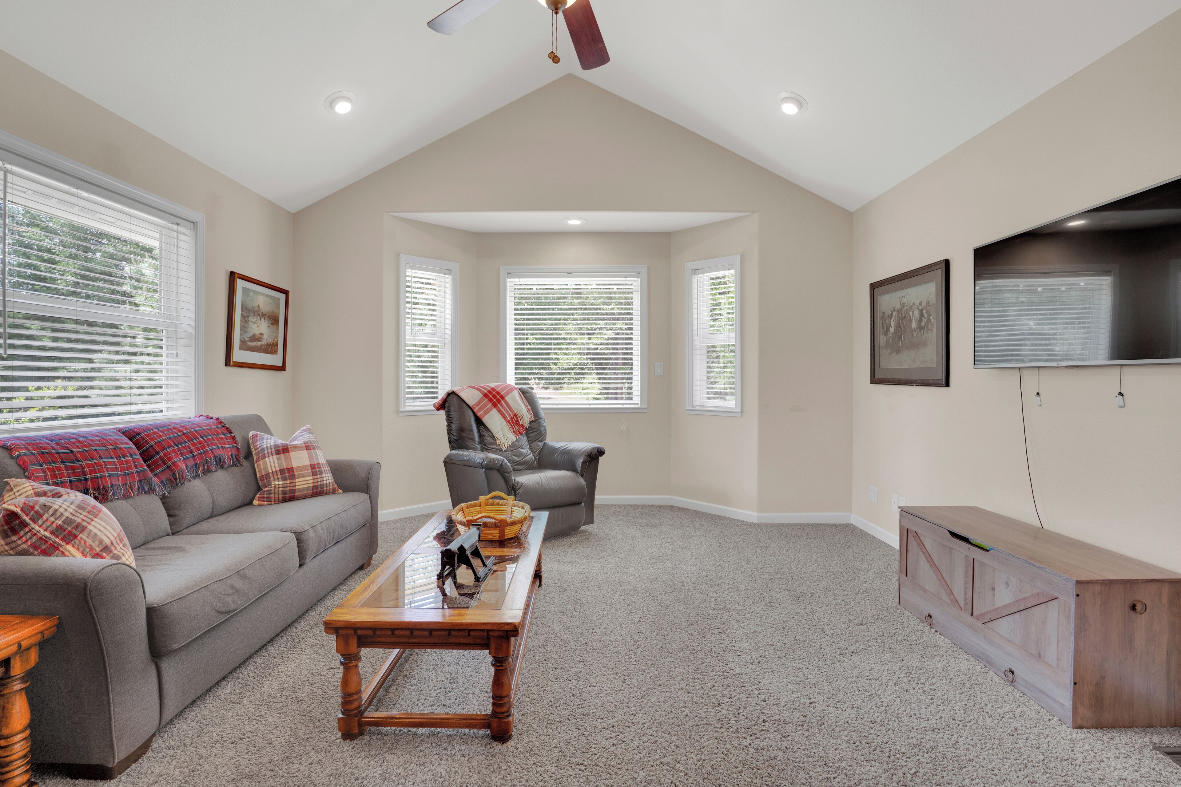 A spacious living room with a high ceiling, a grey sofa with plaid cushions, a wooden coffee table, and a TV on the wall.