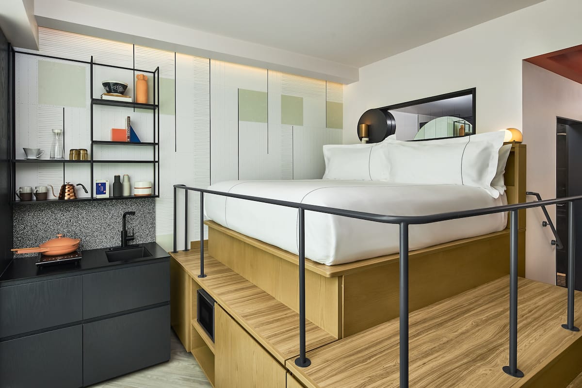 A modern bedroom with a bed, built-in shelves, a small kitchen area, and a mirror.