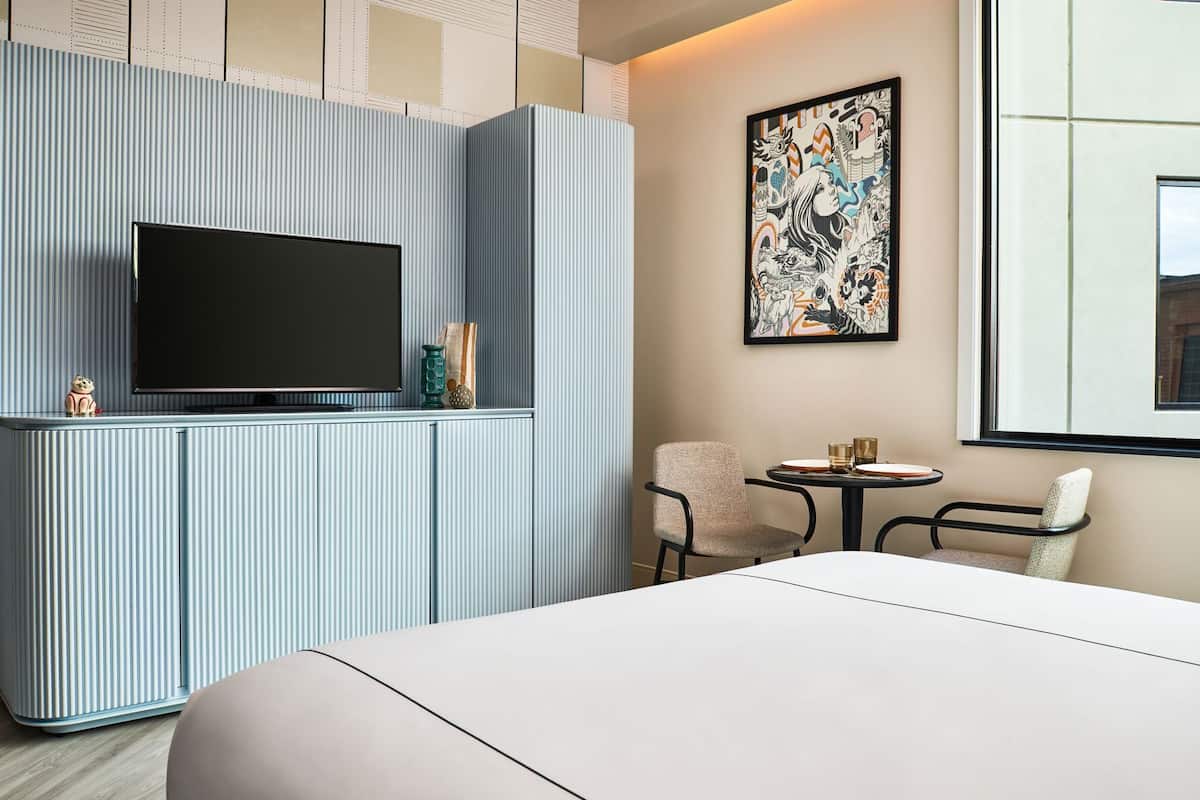 A hotel room with a large bed, a TV on a cabinet, a small table with chairs, and a painting on the wall.