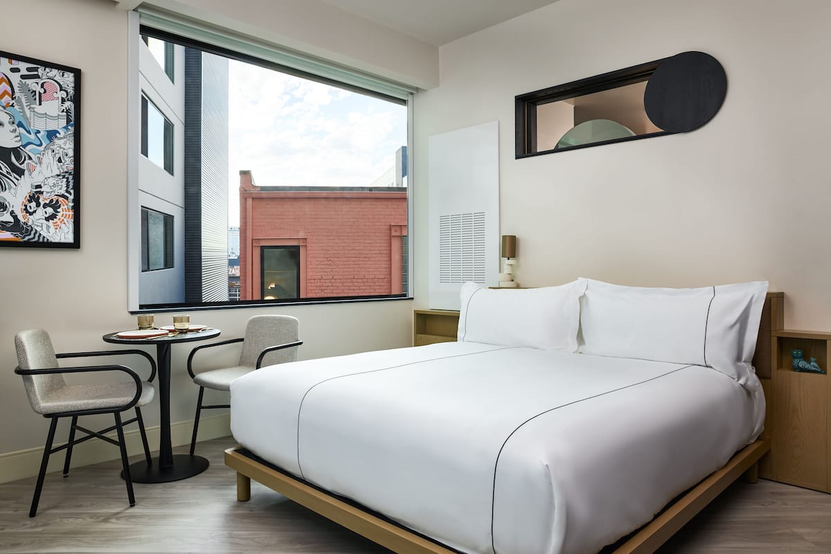 A modern hotel room with a large bed, a small table with two chairs, a window with a view of a building, and a wall-mounted mirror.
