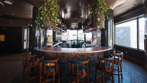 Una zona de bar con mostrador redondo, taburetes de madera, plantas colgantes y un ventanal grande.