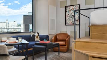 A modern living room with a sofa, a brown leather chair, a dining table with plates and glasses, and a large window with a city view.